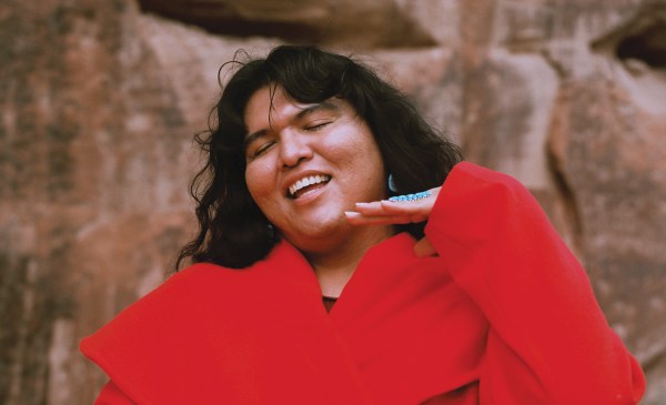 A portrait of charlie amáyá scott, smiling broadly, wearing a red shirt with their hand near their face.