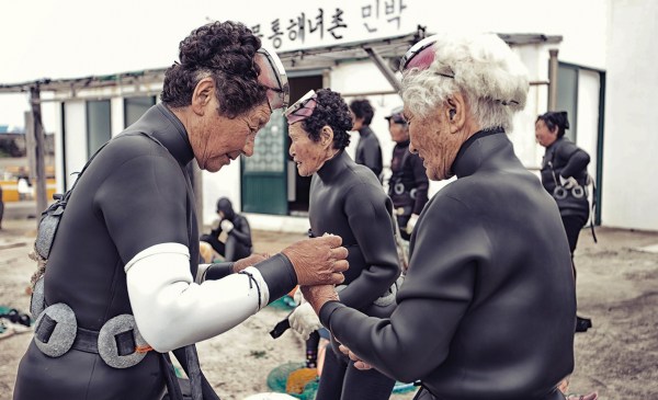 Korean women divers, known as haenyeo, gear up with chest weights (to aid diving), an L-shaped weeding hoe, and a net attached to a flotation device. A wetsuit, goggles, and gloves allow haenyeo divers to stay in the water for between five and six hours, even during the wintertime. Two haenyeo are in central focus in the frame, with a black-haired woman helping a white-haired woman put on gloves.