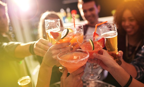 A group of people toast with several colorful cocktails and pints of beer. The people are smiling and the sun is shining.