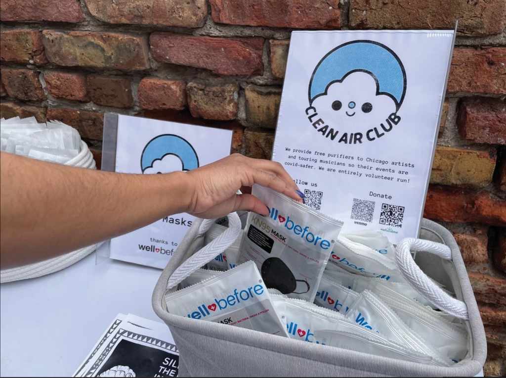 A color photograph of a table at a Clean Air Club event. There is a basket full of maks, and an arm reaches in to select one. 