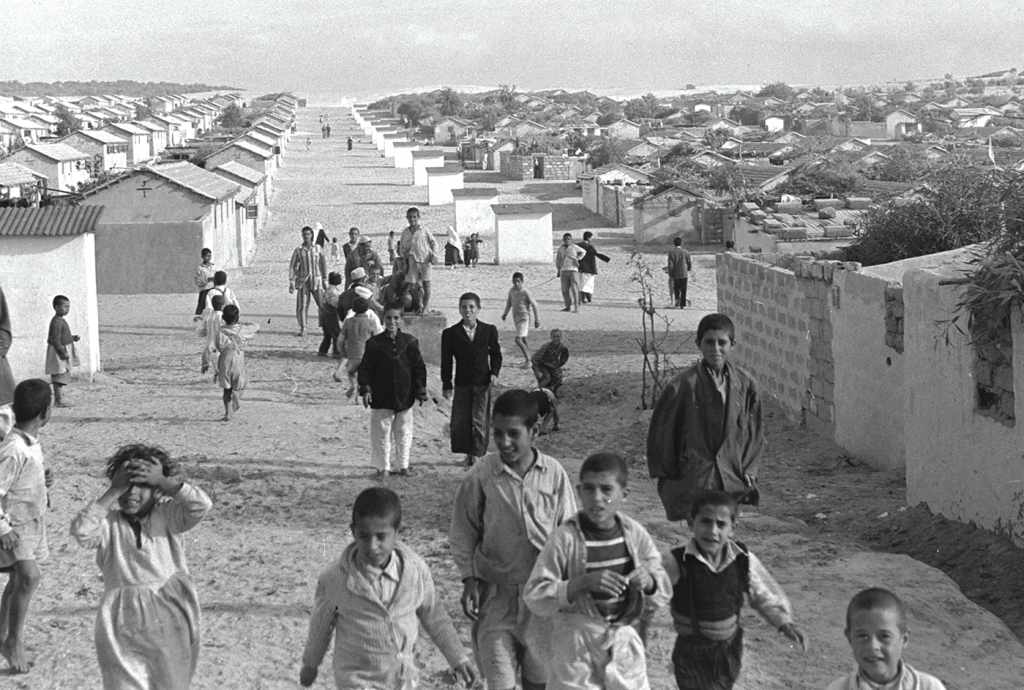 A black and white photograph taken in Gaza City, 1956. A group of around 30 displaced Palestinians, mostly children, walk through a refugee camp in Gaza City during the first Israeli occupation of the Gaza Strip