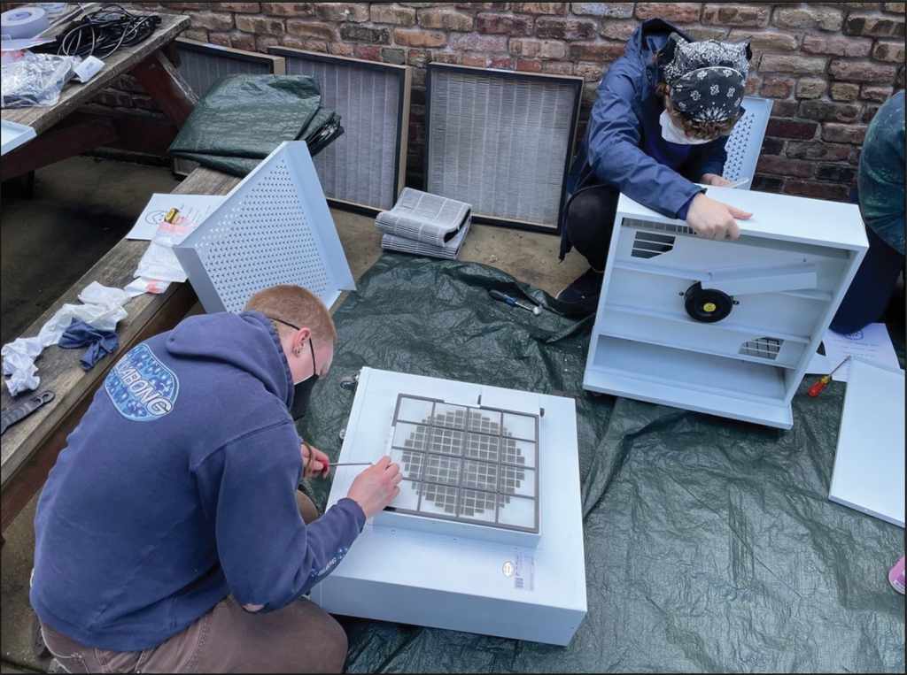 Two masked adults hover over small, white box air purifiers to repair them. 