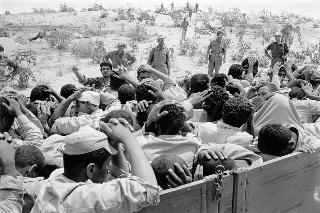 A black and white photo from 1967, during the Naksa, or Six-Day War. A truck bed is full of Palestinian male prisoners with their hands above their heads. Israeli soldiers look on from outside the truck.