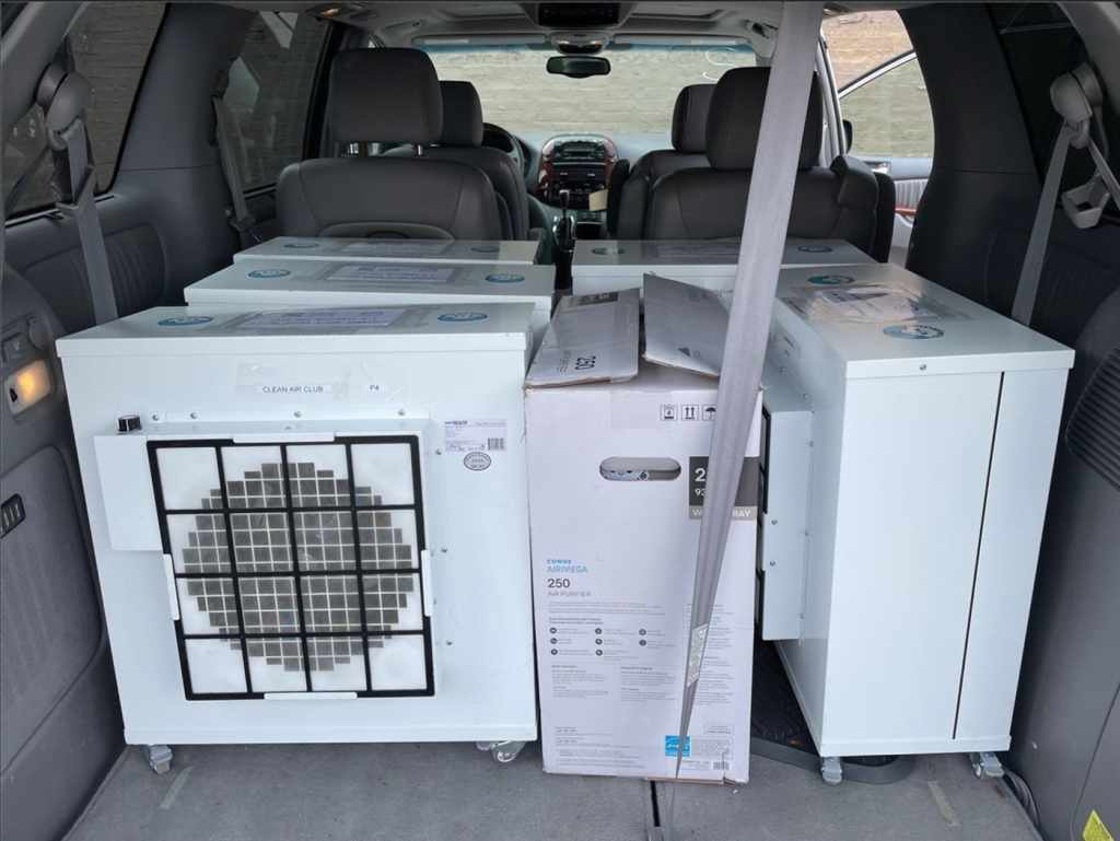 A color photograph from the rear of a van or SUV. The back is filled with the small, white air purifiers to distribute.  