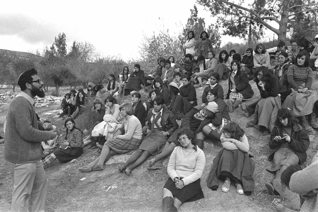 A black and white photograph from December 1975 of Israeli settler leader Benny Katzover speaking to a large group of seated female Jewish settler in the West Bank.