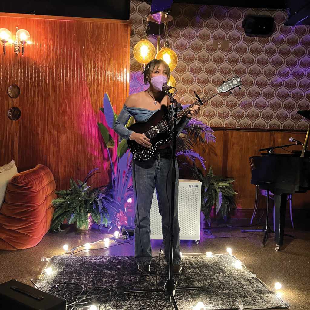 A color photograph from Fruit Salad, the monthly queer open mic. A person holds a guitar and is clearly smiling behind their purple n95 mask.