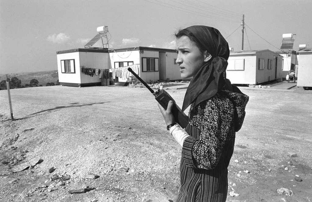 A black and white photograph from 1984. A very young female West Bank settler stands outside her caravan home in the West Bank settlement of Dolev, and uses a walkie-talkie to communicate with a nearby settlers’ council.