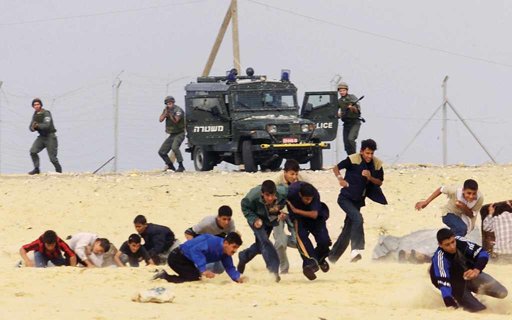 A color photograph from October 24, 2000 features 13 Palestinian male youth in the foreground and an Israeli polic jeep in the background and three Israeli soldiers with automatic weapons. One of them aims at the Palestinians, who had been throwing stones at them.  