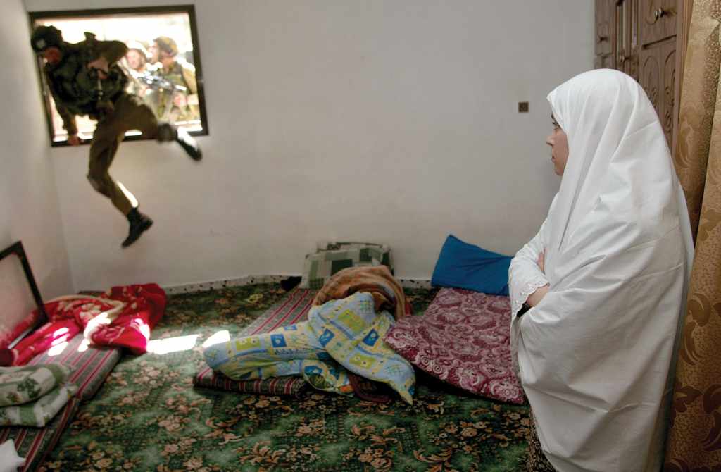 A color photograph from March 2000. A Palestinian woman watches with almost boredom from her room in the West Bank as an Israeli soldier jumps in through the window. More soldiers are behind him. 