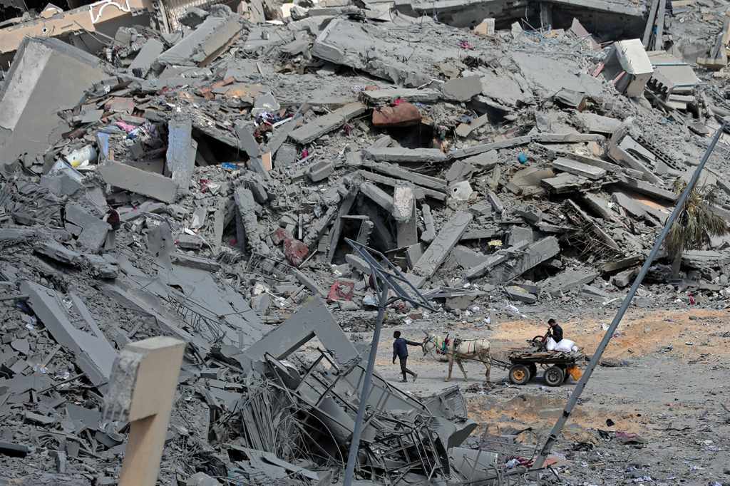 A photo from March 2024 in Gaza. Dwarfed by mountains of grey rubble, of what used to be a city, one Palestinian leads a donkey pulling a cart with another Palestinian seated on top. 