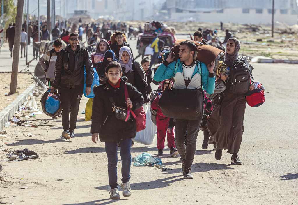 A color photograph from January 2024. A long line of Palestinians travel on foot carrying their belongings leave Khan Yunis. 