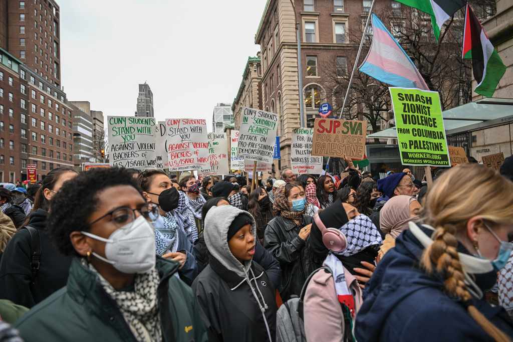 A color photograph from a New York City pro-Palestine protest near Columbia University. Signs from protesters in medical masks and keffiyehs read "CUNY students stand with you," "SJP SVA for a free Palestine," and "Smash Zionist violence, from Columbia to Palestine." 