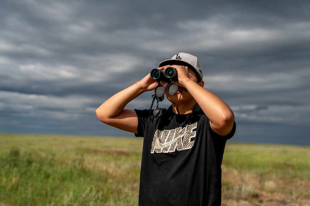 Sage Lone Bear, a Native student dressed in a Nike shirt and baseball cap, stands in an open field and looks out through binoculars. 