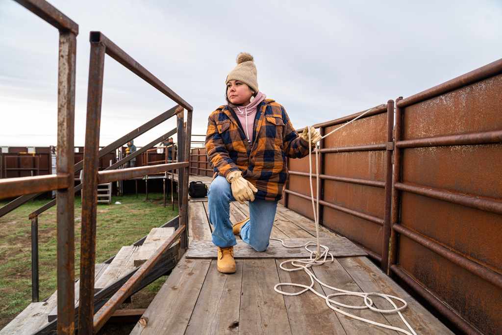 Dawn Thomas, a Native student wearing jeans and a warm jacket, takes a knee on a wooden platform. 