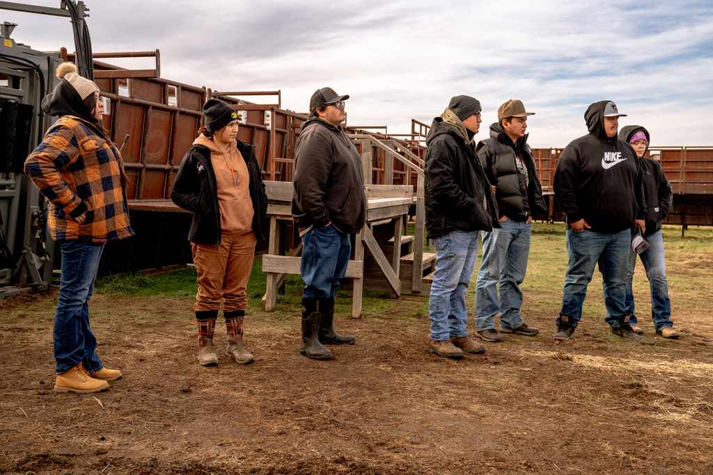 A group of Aaniiih Nakoda College ʔíítaanɔ́ɔ́nʔí/Tatag ́a Buffalo Center students stand near a bison handling area. They are dressed in jeans, boots, warm jackets, and hats. 