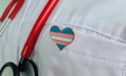 Photo of trans flag colored heart on a doctor's pin.